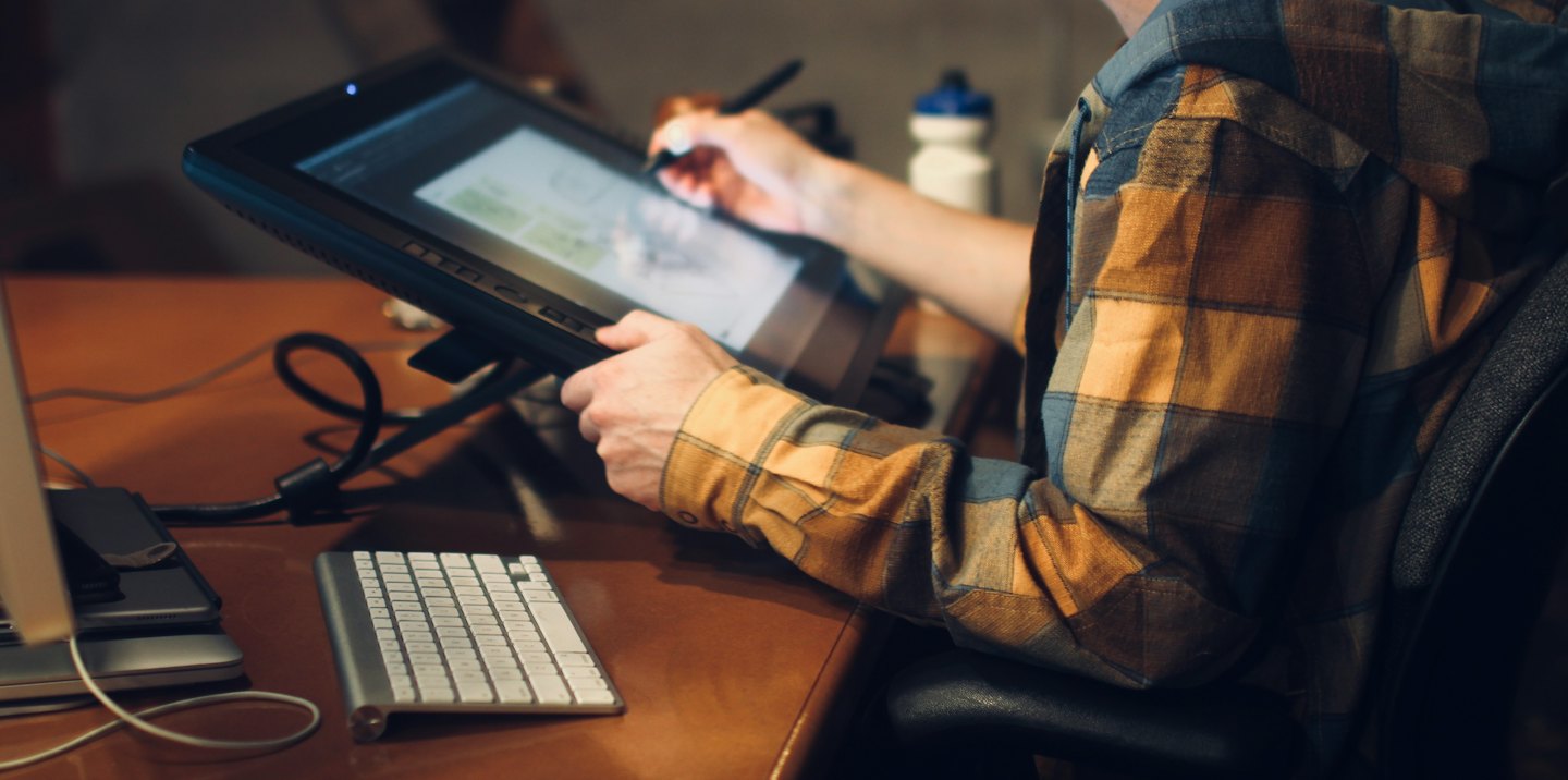 Designer using stylus on drawing tablet at wooden desk.